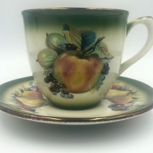 May include: A vintage teacup and saucer set with a green rim and a fruit design. The cup and saucer feature a white background with a green, yellow, red, and purple fruit design.