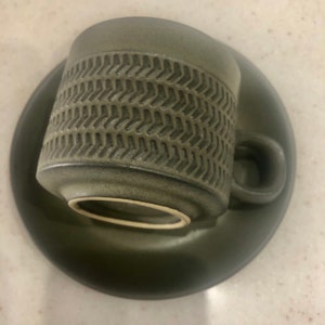 May include: A green ceramic teacup and saucer set with a textured chevron pattern. The cup has a white rim.
