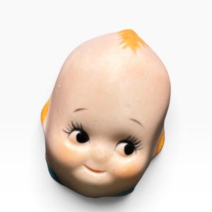 May include: A small ceramic figurine head featuring a pale, smooth complexion, large black eyes with long eyelashes, and a subtle smile. The head has a small tuft of orange hair at the top and orange accents on the sides, with a blue base visible.