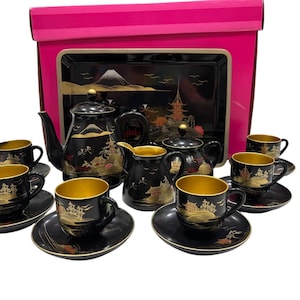 May include: A black and gold Japanese tea set with a teapot, creamer, sugar bowl, cups, and saucers. The set features a landscape design with a mountain and pagoda. The set is displayed with a matching serving tray and a pink box.