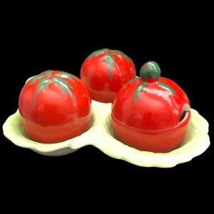 May include: A set of three ceramic tomato-shaped condiment containers with lids. The containers are red with green leaves and are sitting on a green leaf-shaped tray.