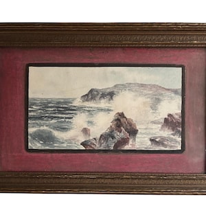 May include: A framed watercolor painting of a seascape. The artwork depicts waves crashing against rocky cliffs under a cloudy sky. The painting is set within a red mat and a decorative gold-toned frame.