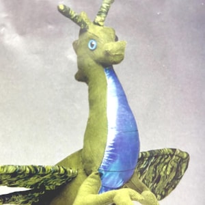 May include: A plush dragon toy featuring an olive green body with a textured surface, a bright blue iridescent underbelly, and two large wings with a dark green and black marbled pattern. The creature has two small horns and light blue eyes.