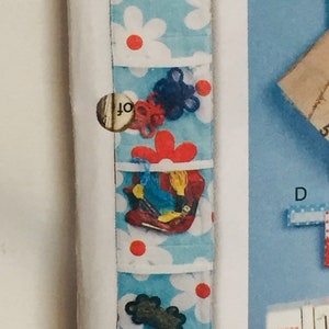 Puede incluir: Un organizador de pared de tela azul y blanco con estampado de margaritas rojas y blancas. El organizador tiene cinco bolsillos y est&aacute; lleno de varios art&iacute;culos, incluyendo hilo, cuerda y un peque&ntilde;o zapato rojo.