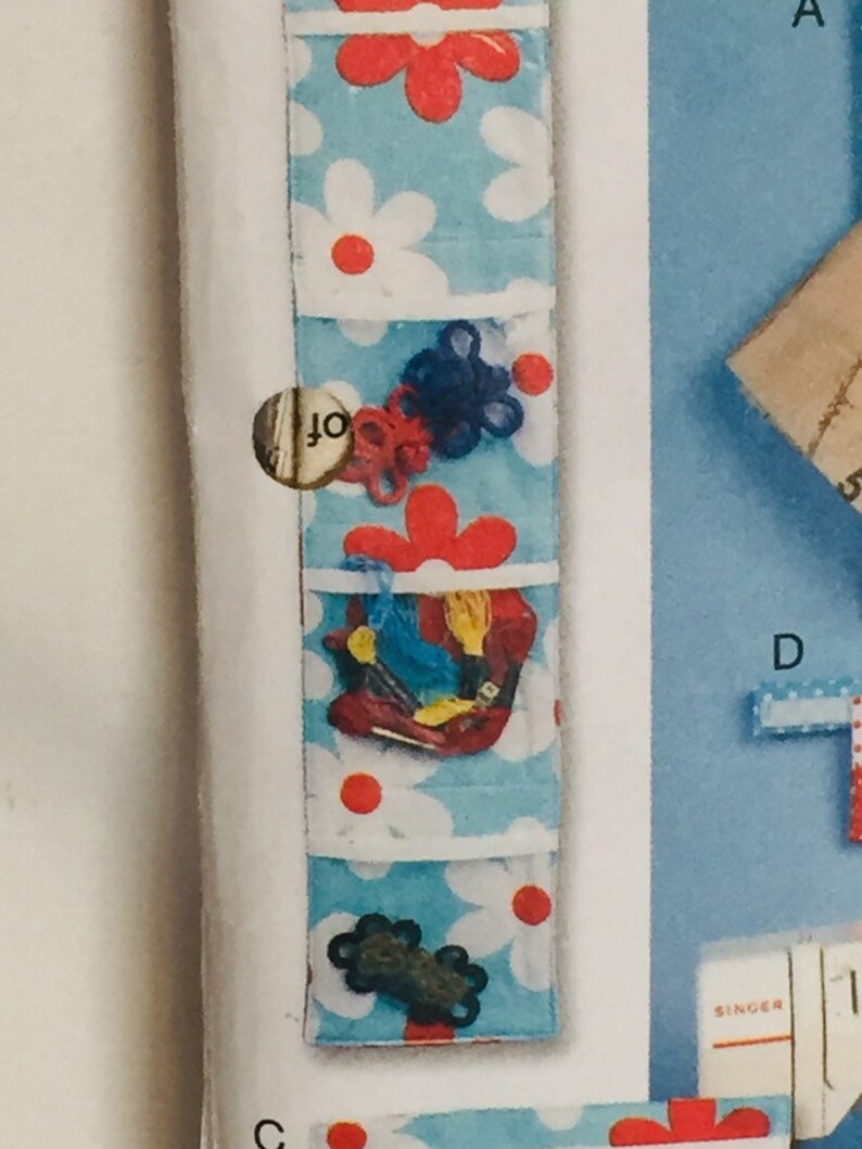 Puede incluir: Un organizador de pared de tela azul y blanco con estampado de margaritas rojas y blancas. El organizador tiene cinco bolsillos y est&aacute; lleno de varios art&iacute;culos, incluyendo hilo, cuerda y un peque&ntilde;o zapato rojo.