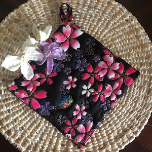 May include: A black quilted hot pad with a pink and white floral pattern. The hot pad has a loop handle and is perfect for protecting surfaces from hot dishes.