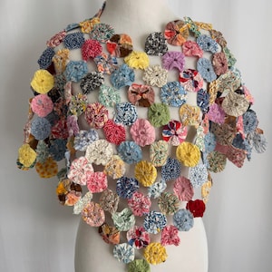 May include: A handmade poncho constructed from numerous circular fabric yo-yos in a variety of colors and patterns. The garment features a lattice-like structure where each small, gathered fabric circle is connected to form a lightweight, open-weave cape.