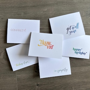 May include: A collection of white greeting cards with various messages in different colors. The messages include "Congrats!", "Thank You", "Get Well Soon", "Happy Birthday", "Thinking of You", and "In Sympathy". The cards are arranged on a wooden surface.