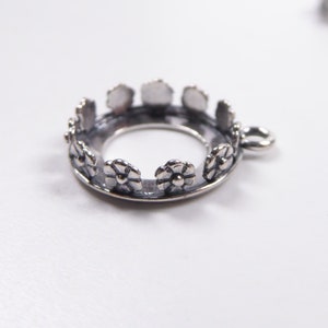 Könnte beinhalten: Silberfarbener Metallanhänger mit einem runden, offenen Zentrum und einem floralen Design am Rand. Der Anhänger hat eine kleine Öse oben zum Anbringen einer Kette.
