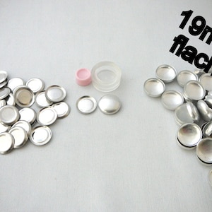 Könnte beinhalten: Eine Ansammlung von silberfarbenen, flachen, runden Metallkomponenten, möglicherweise für Bastelarbeiten oder Schmuckherstellung. Das Bild zeigt auch eine kleine rosa und transparente Kunststoffkomponente. Der Text im Bild lautet "19mm flach".