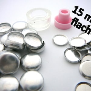 Könnte beinhalten: Eine Ansammlung von silberfarbenen Metall-Cabochon-Fassungen mit flachen Rückseiten und passenden runden Einsätzen. Ein klarer Kunststoffbehälter und ein rosa Kunststoffwerkzeug sind ebenfalls vorhanden. Der Text "15 mm flach" ist sichtbar.