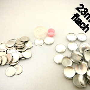 Könnte beinhalten: Eine Ansammlung von silberfarbenen, flachen, runden Metallkomponenten, zusammen mit einem transparenten Kunststoffbehälter und einem kleinen rosa Deckel. Der Text "23mm flach" ist in Schwarz sichtbar. Diese Komponenten scheinen Bastelbedarf zu sein.