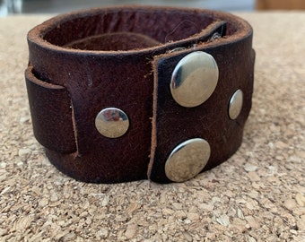 May include: A wide, dark brown leather wrist cuff with silver-colored snap closures. The cuff is thick and appears to be made of distressed leather. The cuff is designed to wrap around the wrist and fasten with snaps.