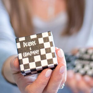 May include: A small, square gift box with a black and white checkerboard pattern. The text "Deirdre & Company" is printed in white on the top of the box. The box is held in a person's hand.