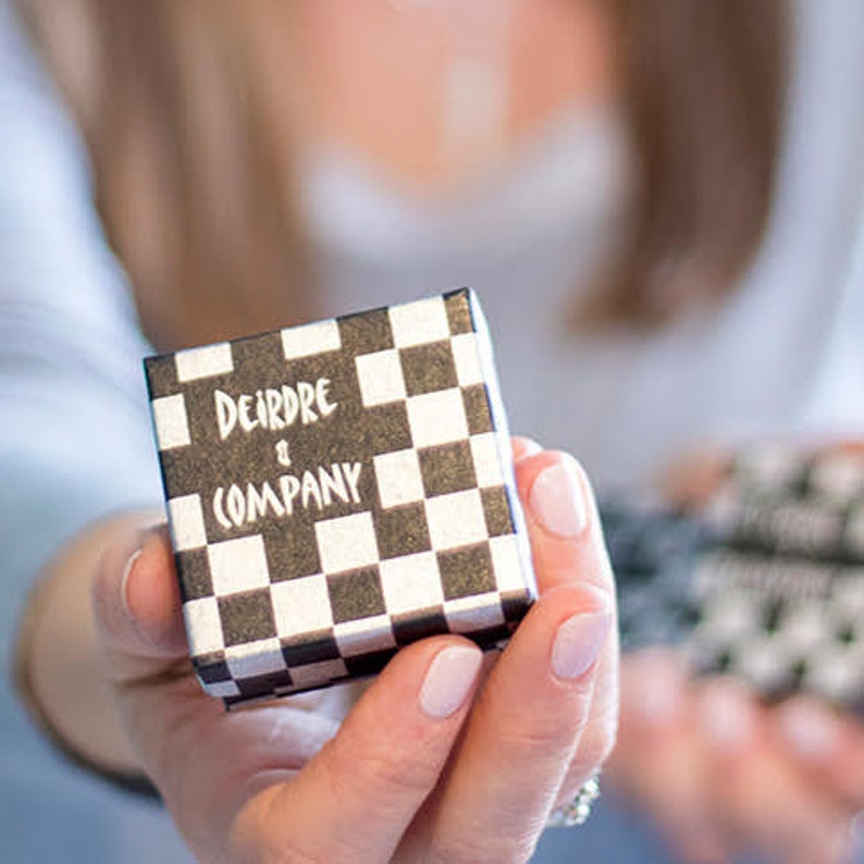 May include: A small, square gift box with a black and white checkerboard pattern. The text "Deirdre & Company" is printed in white on the top of the box. The box is held in a person's hand.