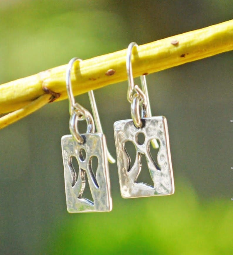 May include: A pair of silver dangle earrings. Each earring features a rectangular pendant with a cut-out angel design. The earrings are suspended from a simple hook. The background is a blurred green and yellow.