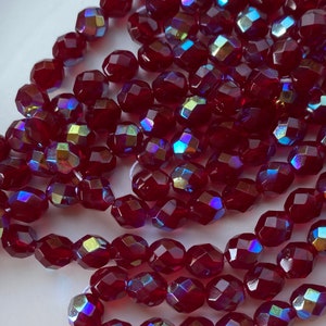 May include: Close-up of faceted, dark red beads with iridescent highlights. The beads are strung together, creating multiple strands. The beads have a geometric, multi-faceted design, reflecting light and creating a sparkling effect. Ideal for jewelry making.