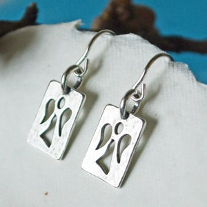 May include: A pair of silver-toned dangle earrings. Each earring features a rectangular tag with a cut-out angel design. The earrings have a hammered texture and are suspended from curved ear wires. The background is a soft white and blue.