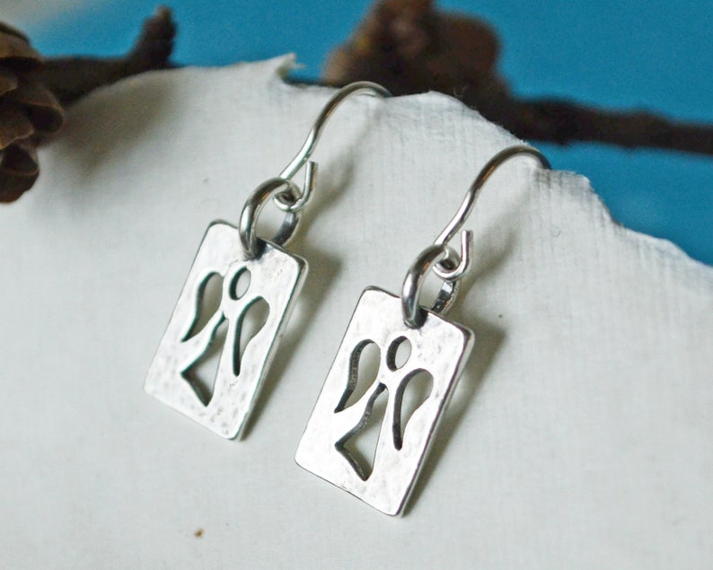 May include: A pair of silver-toned dangle earrings. Each earring features a rectangular tag with a cut-out angel design. The earrings have a hammered texture and are suspended from curved ear wires. The background is a soft white and blue.