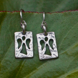 May include: A pair of silver-toned earrings. Each earring features a rectangular pendant with a textured surface and a cut-out angel design. The earrings hang from curved ear wires. The background is a green leaf.