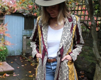 Könnte beinhalten: Eine Frau trägt eine lange, gemusterte Jacke mit einem geometrischen Muster in Gelb, Rot und Schwarz. Sie trägt auch eine blaue Jeans, ein weißes T-Shirt, einen braunen Gürtel und schwarze Stiefel.