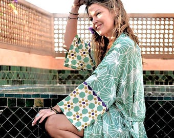 Op de afbeelding: Een vrouw draagt een groene en witte kimono met geometrisch patroon en een gouden en groene geometrische rand. De kimono heeft een strikband en wordt gedragen over een wit topje.
