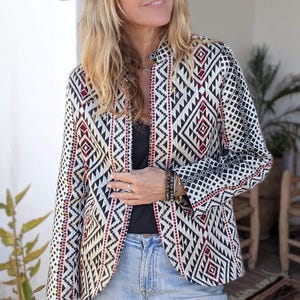 May include: A patterned jacket with a black, white, and red geometric design. The jacket is worn with a black top, blue jeans, and a green hat. The jacket's pattern is a striking visual element.