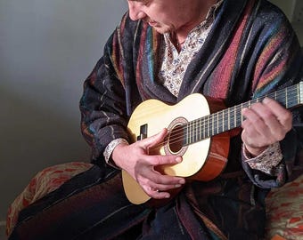 Puede incluir: Una persona tocando una pequeña guitarra acústica. La guitarra es de color marrón claro con una boca redonda. La persona lleva una bata estampada con rayas rojas, azules y verdes. Se ve un libro titulado "Tíbet".