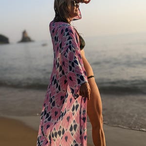 Könnte beinhalten: Eine Frau trägt einen pink- und blaugemusterten Kimono mit einem schwarzen Bikini-Oberteil und -Unterteil. Sie steht an einem Sandstrand mit dem Meer im Hintergrund.