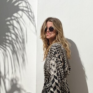 May include: A woman wearing round sunglasses and a black and white geometric print kimono-style dress. The dress has a long sleeve and a tie belt. The woman is standing in front of a white wall with a shadow of a palm tree.