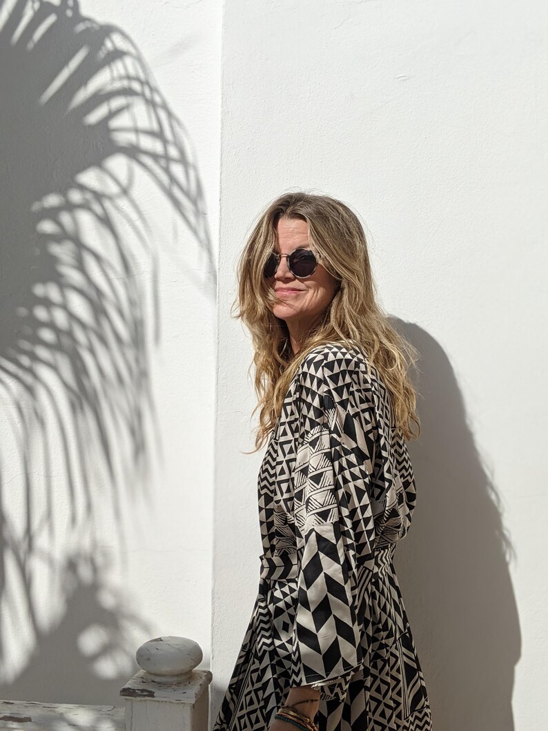 May include: A woman wearing round sunglasses and a black and white geometric print kimono-style dress. The dress has a long sleeve and a tie belt. The woman is standing in front of a white wall with a shadow of a palm tree.