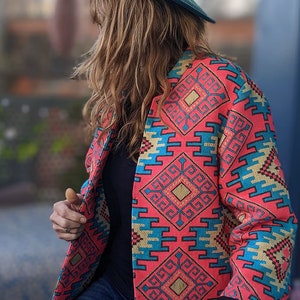 May include: A teal fedora hat with a brown leather band and buckle. A coral-coloured jacket with a geometric pattern in teal and gold is worn over a black top and blue jeans. The jacket's design is a striking visual element.