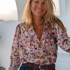 Könnte beinhalten: Eine Frau trägt eine weiße, rosa und lila gemusterte Bluse mit einem dunkelblauen Gürtel und einer dunkelblauen Jeans. Sie lächelt und schaut in die Kamera.