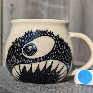 May include: Cream-colored ceramic mug featuring a black and white monster design. The monster has a large eye, sharp teeth, and a textured, spiky outline. The mug has a curved handle and a blue sticker with the text "M 301".