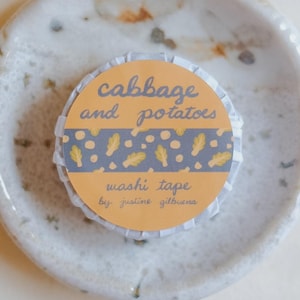 May include: A roll of washi tape with the text "cabbage and potatoes" in a decorative font. The tape has a yellow and blue design with feather and dot patterns. The tape is on a small, speckled ceramic dish.