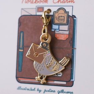 May include: Gold-tone metal notebook charm featuring a gray bird carrying a brown envelope. The charm is attached to a gold-tone metal chain. The charm is displayed on a white card with the text "Notebook Charm" and "illustrated by justine gilbuena".