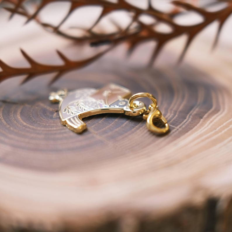 May include: A gold-colored metal charm with a detailed design of a bird with floral accents. The charm has a loop for attaching to a keychain or necklace.