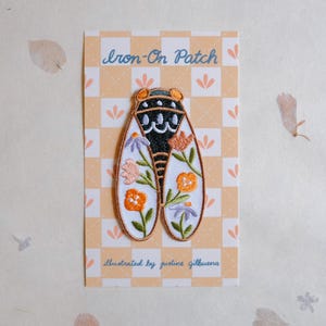 May include: An iron-on patch with a cicada design, adorned with orange, purple, and pink floral details. The patch is presented on a card with the text "Iron-On Patch" and "illustrated by justine gilbuena". The background features a light orange checkered pattern.