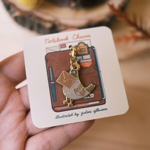 May include: Gold-tone metal charm featuring a bird carrying a brown envelope. The charm is attached to a gold chain and is labeled "Notebook Charm" and "illustrated by justine gilbuena".
