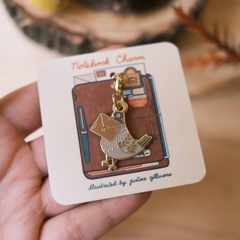 May include: Gold-tone metal charm featuring a bird carrying a brown envelope. The charm is attached to a gold chain and is labeled "Notebook Charm" and "illustrated by justine gilbuena".