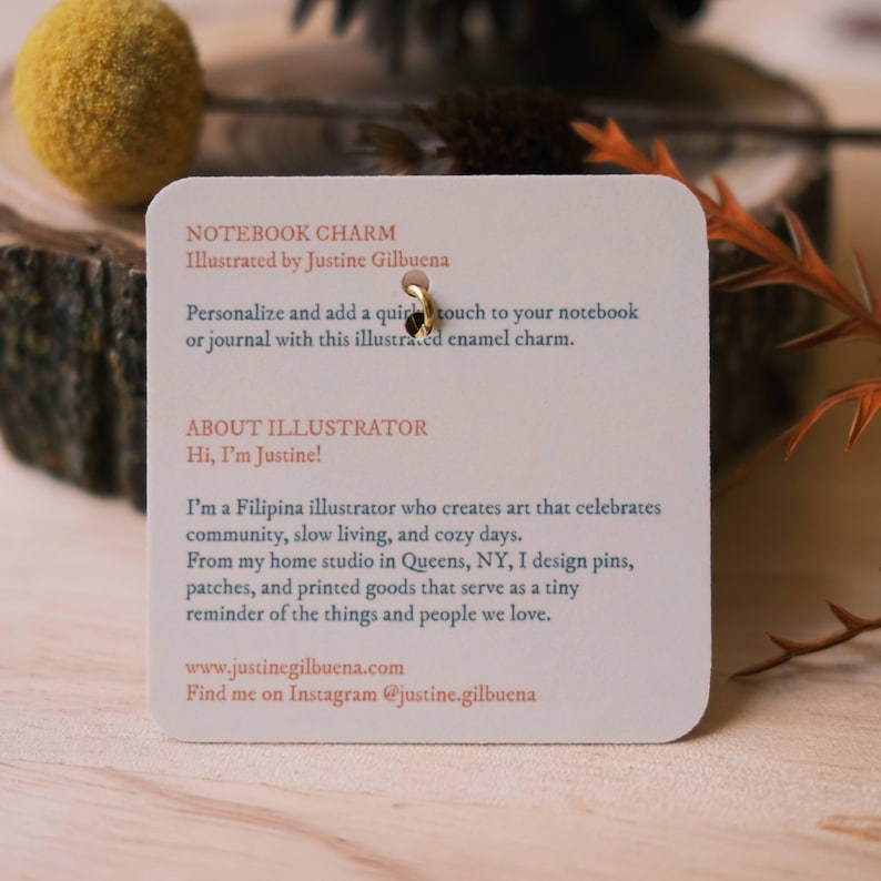 May include: A white card with the text "Notebook Charm" and "Illustrated by Justine Gilbuena". The card also includes information about the illustrator, Justine Gilbuena, and her work. The card is attached to a gold metal ring.