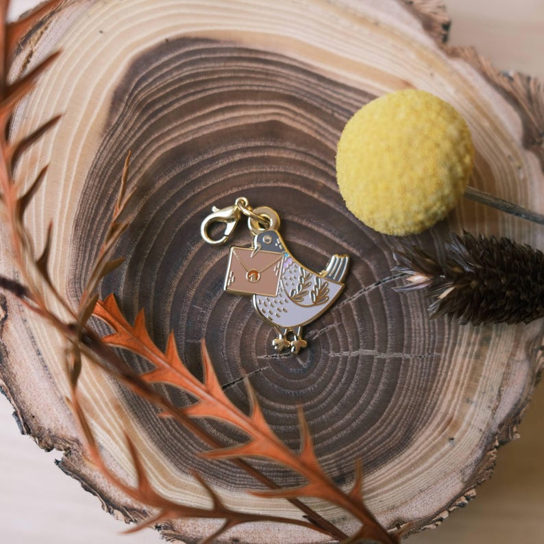 May include: A gold-tone charm featuring a white dove carrying a brown envelope. The charm has a gold-tone clasp for attaching to a necklace, bracelet, or keychain.