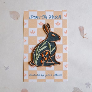 May include: An iron-on patch featuring a stylized rabbit design. The rabbit is dark teal with brown ears and is decorated with green leaves and orange berries. The patch is on a card with the text "Iron-On Patch" and "illustrated by justine gilbuena".