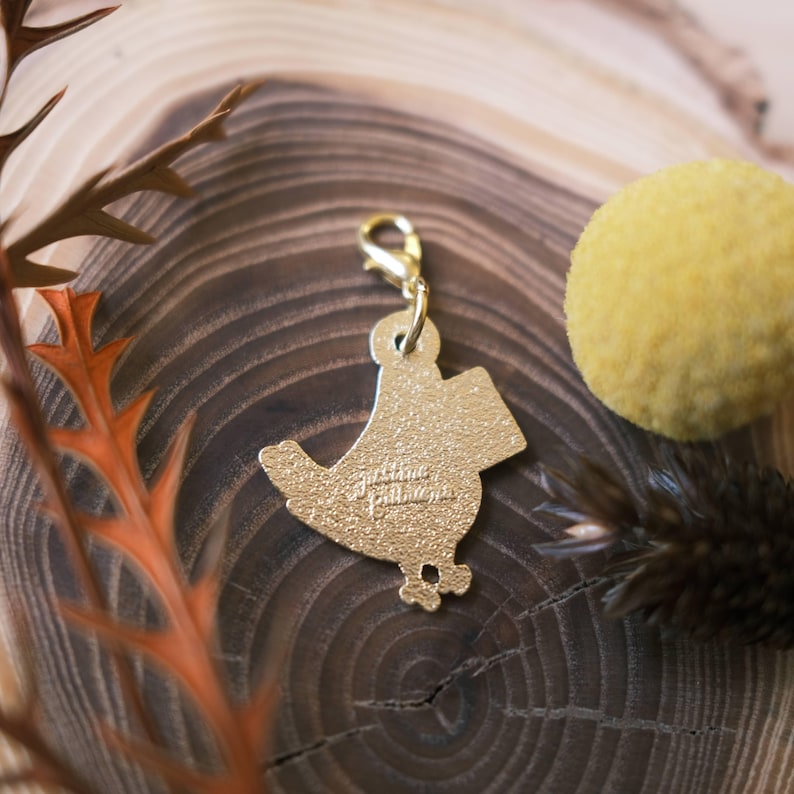 May include: Gold-colored metal charm shaped like a bird with the text "Justine Bailey" on the back. The charm has a lobster clasp.