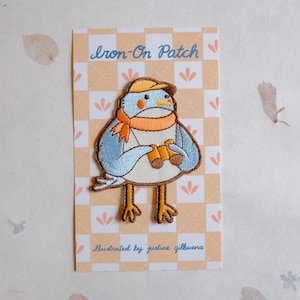 May include: An embroidered iron-on patch featuring a blue bird wearing a hat, scarf, and holding binoculars. The patch is on a card with the text "Iron-On Patch" and "illustrated by justine gilbuena". The background is a light orange and white checkered pattern.