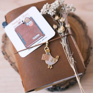 May include: A brown leather journal with a gold bird charm attached. The bird is holding a small envelope in its beak. The journal has a white card attached to it with the text "Notebook Charm".