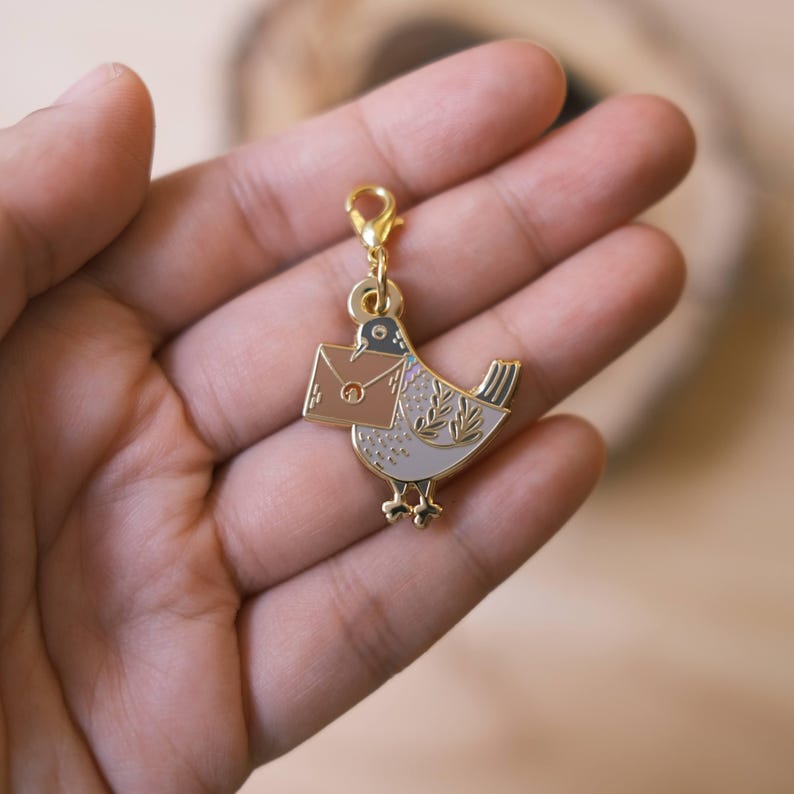May include: Gold-tone metal charm featuring a white and brown bird carrying a brown envelope. The charm has a lobster clasp.