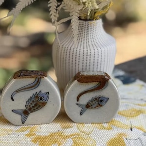 May include: Ceramic salt and pepper shakers featuring a fish design. The shakers are white with brown and blue fish illustrations. A white vase with a dotted pattern and a bouquet of dried flowers is in the background.
