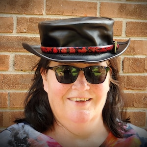 May include: A woman wearing a black leather hat with a red and black snake skin band. She is wearing sunglasses and has a smile on her face.