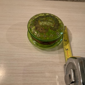 May include: A green glass trinket box with a gold floral design and a gold inscription. The box is approximately 2 inches in diameter.